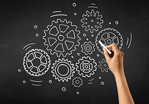 Female hand holding white chalk in front of a blackboard with gears drawn on it